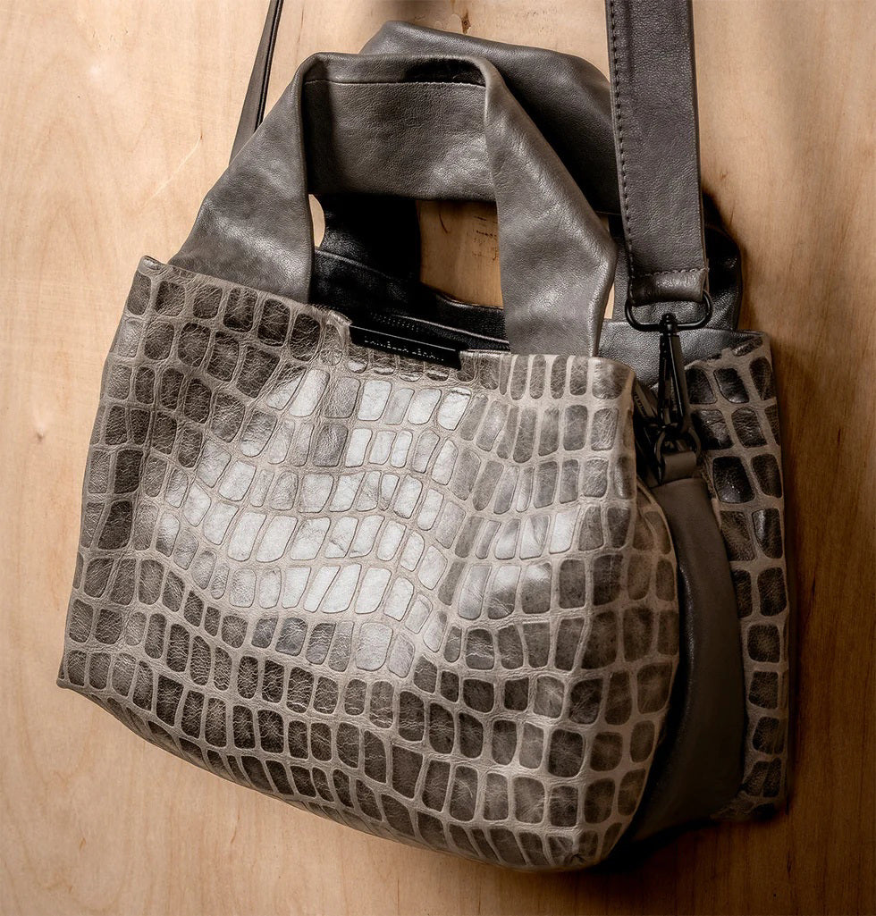 The DANIELLA LEHAVI Eveline Satchel in Grey Mosaic features a croco-inspired textured leather design with short handles and a long shoulder strap, epitomizing quiet luxury as it hangs gracefully against a wooden background.