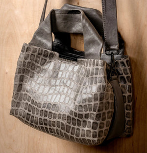 The DANIELLA LEHAVI Eveline Satchel in Grey Mosaic features a croco-inspired textured leather design with short handles and a long shoulder strap, epitomizing quiet luxury as it hangs gracefully against a wooden background.
