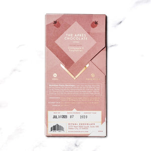 A rectangular chocolate bar with a pinkish wrapper adorned with minimalist illustrations of raspberries and abstract shapes. The packaging reads "RITUAL CHOCOLATE - THE APRES CHOCOLATE BAR 70%, Sparkling Wine Infused Chocolate with Dried Raspberries," 2.12 oz (60g).