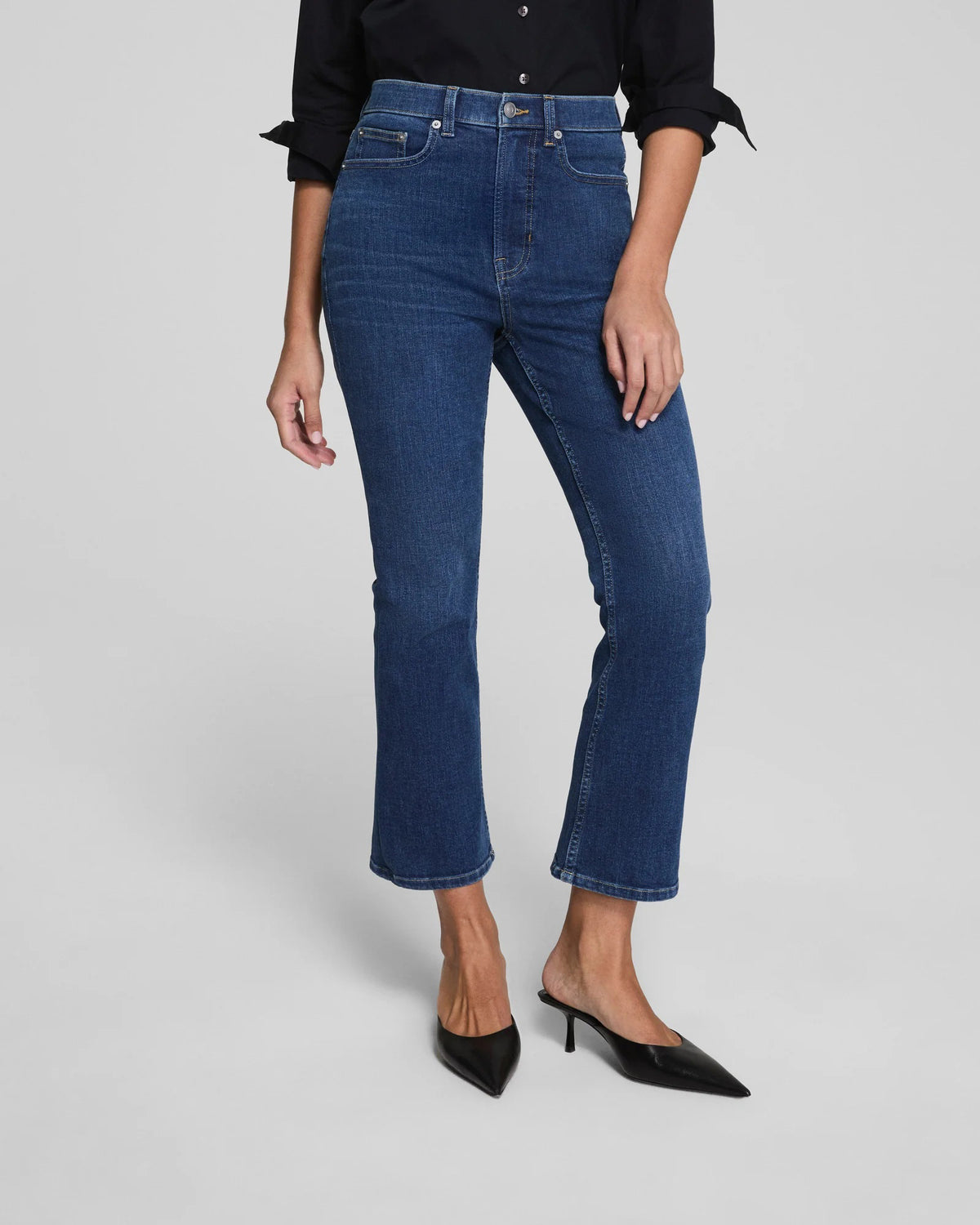 A woman with her hair pulled back stands against a plain background wearing SPANX Redefine Kick Flare Jeans in Medium Indigo, a black button-up shirt, black pointed-toe heels, and carries a black shoulder bag with one hand in her pocket.
