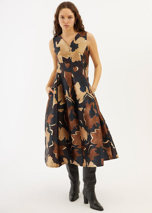 A woman stands with her hands in the pockets of the PARVATI DRESS by ALYKI LABORATORIO, a sleeveless, V-neck, calf-length dress featuring a brown and black abstract floral pattern, styled with black knee-high boots.