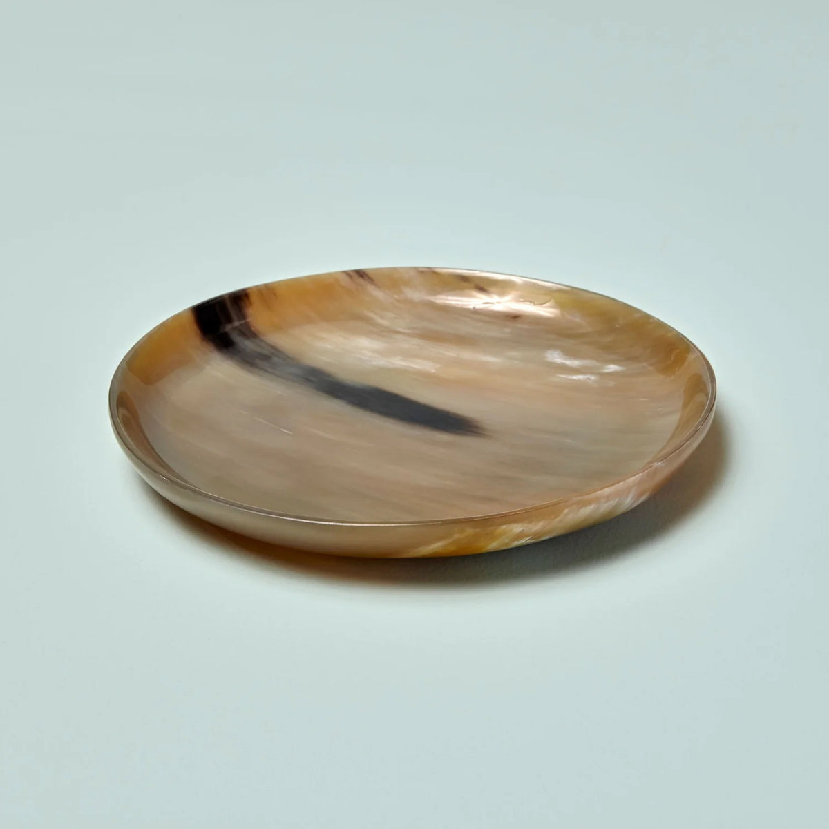 The SPLATTER HORN PLATE MEDIUM by BE HOME, INC is a round, shallow dish with a smooth and polished surface, ethically crafted from water buffalo horn. This handmade marvel from Vietnam displays a natural, marbled pattern in shades of beige, brown, and black. It rests on a flat surface with simple, elegant contours.
