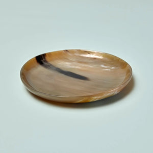 The SPLATTER HORN PLATE MEDIUM by BE HOME, INC is a round, shallow dish handcrafted in Vietnam from polished water buffalo horn, showcasing a marbled pattern of light brown, cream, and dark brown tones.