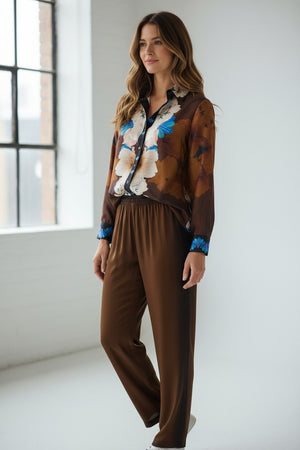 A woman stands indoors by a window, smiling as she looks to the side. She wears the FRANCO FERRARI Long Sleeve Silk Blouse, featuring a vibrant floral print in brown, white, and blue patterns, paired with black high-waisted pants. One hand is in her pocket.