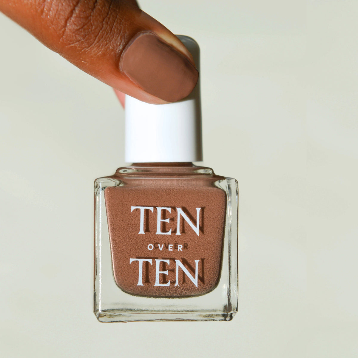 A hand with TEN OVER TEN's vegan WARREN NAIL POLISH in taupe wears a shiny gold ring on the middle finger and three gold bangles, set against a sunlit stone background.
