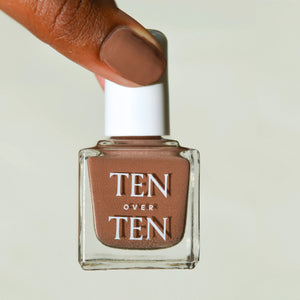 A hand with TEN OVER TEN's vegan WARREN NAIL POLISH in taupe wears a shiny gold ring on the middle finger and three gold bangles, set against a sunlit stone background.