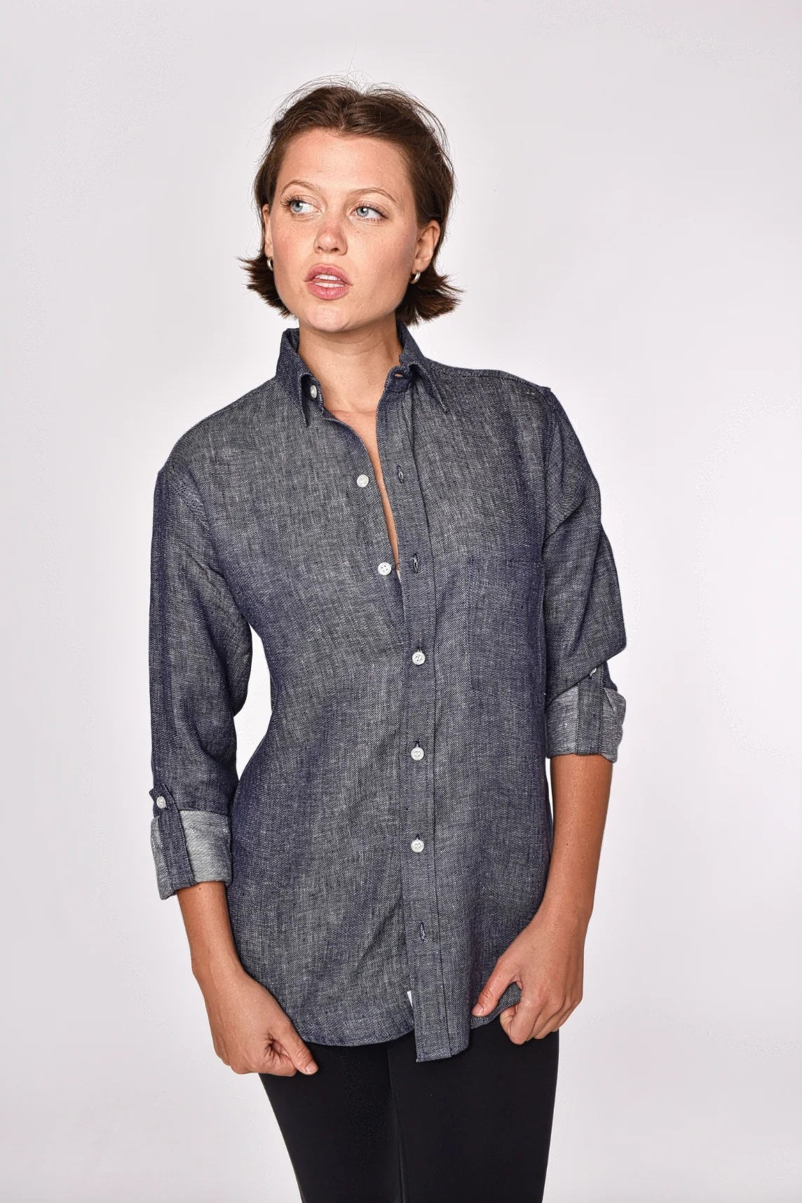 A person with short brown hair, dressed in the EMILY PHILLIPS - THE LINEN DENIM SHIRT—a long-sleeve, button-up gray shirt from EMILY PHILLIPS crafted from breathable linen fabric—and black pants stands against a plain light background, looking slightly to the left with a neutral expression.