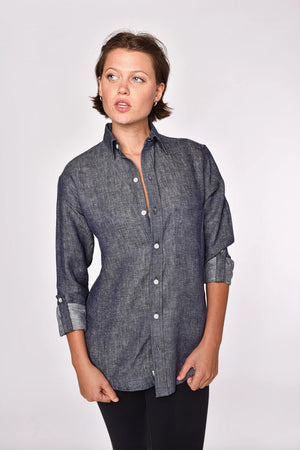 A person with short brown hair, dressed in the EMILY PHILLIPS - THE LINEN DENIM SHIRT—a long-sleeve, button-up gray shirt from EMILY PHILLIPS crafted from breathable linen fabric—and black pants stands against a plain light background, looking slightly to the left with a neutral expression.