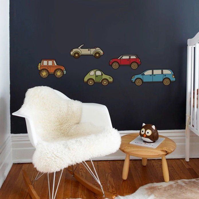 A child's room featuring a white crib-like bed, blue chair, and carpet showcasing a brown, white, and orange circular pattern. SIMPLE SHAPES - CAR REMOVEABLE WALL DECALS by SIMPLE SHAPES in various colors are adhered to the wall above the bed. A blue toy car is positioned on the floor near the bed.