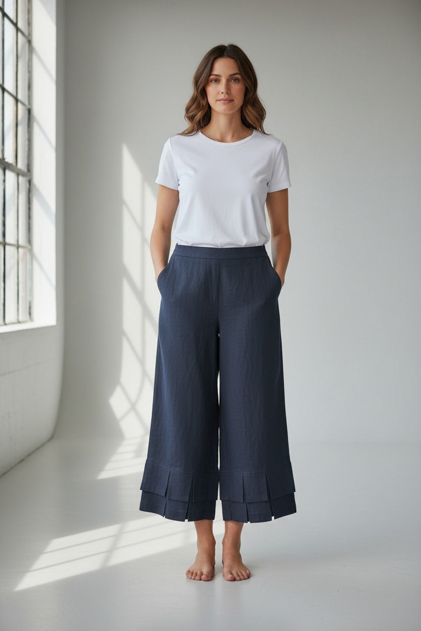 A woman stands barefoot indoors, wearing a white t-shirt and the MEIMEIJ Linen Pull On Pant with Ruffle Cuff by MEIMEIJ in navy blue. She looks at the camera with her hands in her pockets, illuminated by light streaming from large windows to her left.
