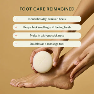 A person uses KATE MCLEOD’s THE FOOT STONE SOLID FOOT MOISTURIZER on their foot, holding the off-white round bar with one hand while the other rests on their ankle. The solid beige background emphasizes care for dry, cracked heels.