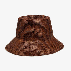 A woman with long, wavy blonde hair wears the WYETH HATS Tali Raffia Bucket Hat in Chocolate by WYETH and a white sleeveless top, looking slightly to the side against a plain light background.