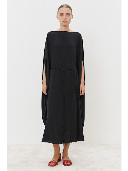 Against a white backdrop, the individual wears a MEIMEIJ Silk Blend Dress with dramatic cape-like sleeves. Their long hair flows down the elegant black gown&#39;s cinched waist, complemented by striking red sandals.