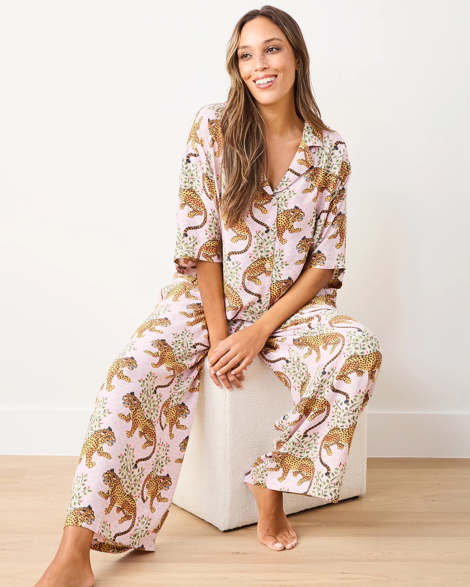 A person on a wooden floor wears the PRINTFRESH - Satin Bagheera Wildest Dream Pajama Set, made of eco-friendly viscose with a leopard print. The loose-fitting top and pants flow gently as they face away, presenting the back of their outfit against a plain, light-colored wall.