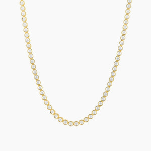 The ELLA STEIN - DIAMOND ESSENTIAL TENNIS NECKLACE by ELLA STEIN is a gold-plated piece adorned with a continuous row of evenly spaced round, white gemstones along the chain, beautifully displayed on a white background. This elegant necklace blends classic design with the lasting durability of stainless steel.