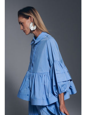 A woman with straight, light brown hair wears the MILVA MI - COTTON BLOUSE IN AZZURO BLUE by MILVA, featuring voluminous sleeves and a peplum hem, paired with bold white earrings against a plain gray background.