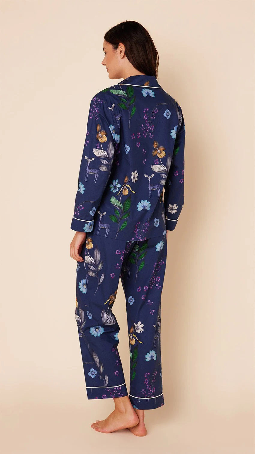 A woman stands barefoot against a plain background, wearing the CAT&#39;S PAJAMAS - Deerly Luxe Pima Cotton Pajamas by CAT&#39;S PAJAMAS. The navy blue pajama set features a colorful floral pattern and is crafted from ultra-soft pima cotton. It includes a long-sleeved button-up shirt and matching pants, both adorned with white piping along the edges.