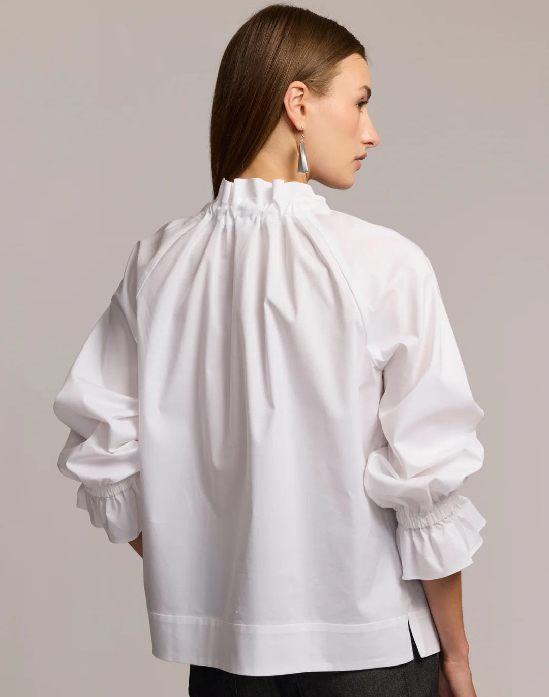 A woman with long brown hair models the HINSON WU ALINA LONG SLEEVE SHIRT, a bright blue blouse with a bow neck and ruffled sleeves, paired with dark pants. She stands against a light background, looking at the camera.