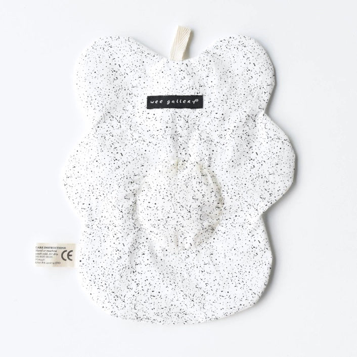 The WEE GALLERY - EGG CRINKLE TOY by WEE GALLERY is a white baby crinkle toy in the shape of a flower with a yellow circle at the center that features a smiling face with closed eyes. Made from organic cotton, it comes with a brown tag inscribed "Baby Crinkle Toy - Organic Crinkle Toy." The toy is displayed hanging on a white hook.