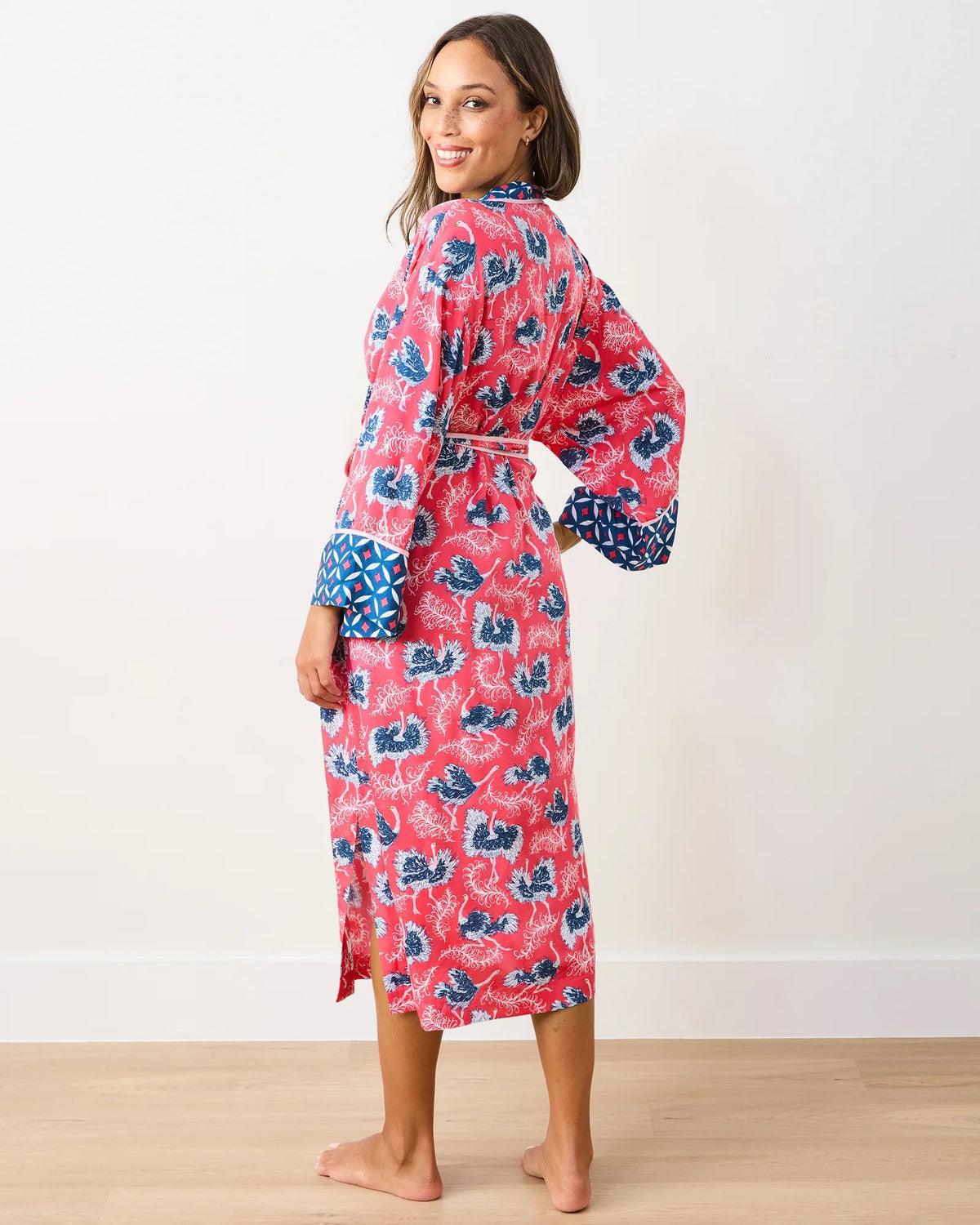 A smiling person, barefoot on wooden flooring against a white wall, wears the vibrant PRINTFRESH - SATIN ROBE OSTRICH DANCE. This eco satin robe has blue and pink floral patterns, a geometric trim, and a waist tie for comfort and style.