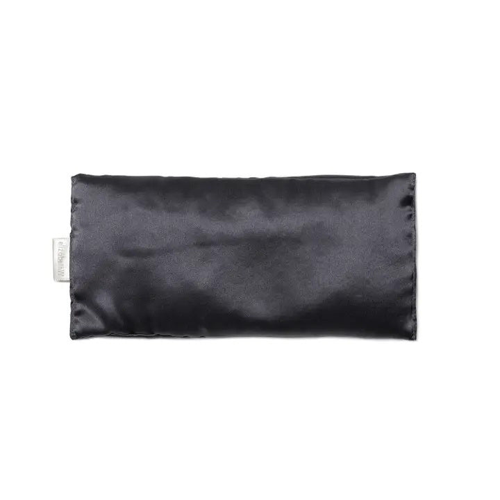 A rectangular, slate-colored satin pillow from E Wightman &amp; Co is shown. The SILK EYE PILLOW - SLATE has a smooth, shiny finish and a small white tag on one side. Its sleek design suggests it could be used for comfort, relaxation, or as a decorative accessory; it could even double as an elegant silk-case eye pillow for ultimate luxury.