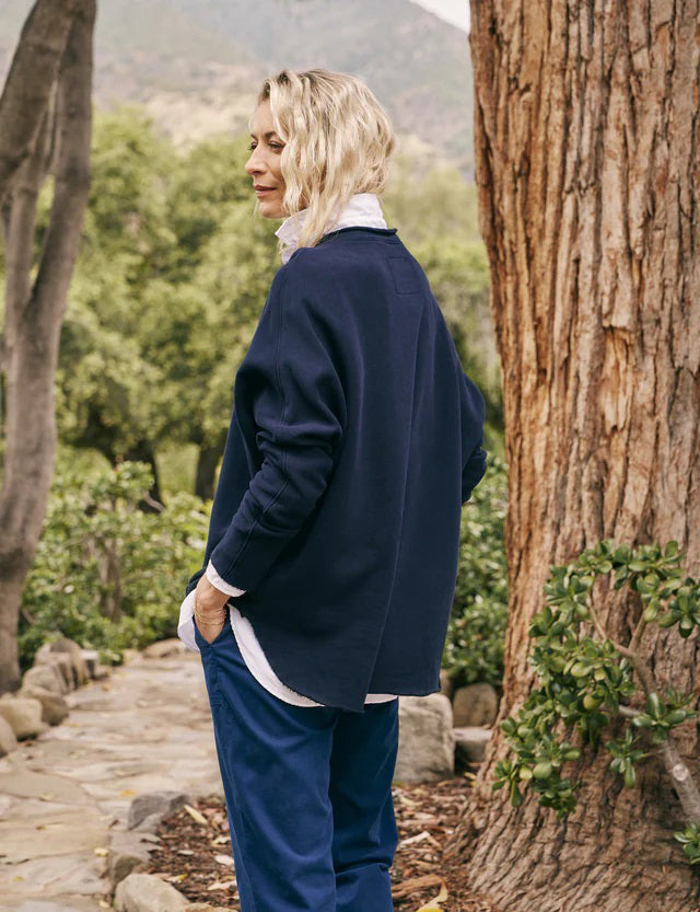 A person with long, wavy blonde hair is standing outdoors near a large tree, wearing the FRANK &amp; EILEEN ANNA LONG-SLEEVE CAPELET in navy over a white shirt and blue pants. The background showcases green foliage and a stone path. The person is smiling and gazing to the side.