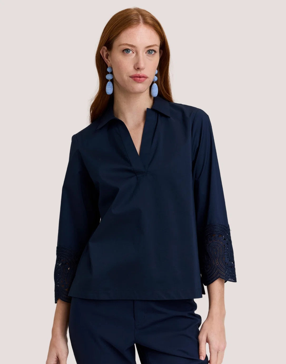 A woman with long red hair wears the HINSON WU MONA Bracelet Sleeve Stretch Polished Cotton Lace Top in navy—a comfortable, stretch cotton blouse featuring three-quarter sleeves with lace accents—paired with navy pants. She accessorizes with blue statement earrings and stands against a plain light background.