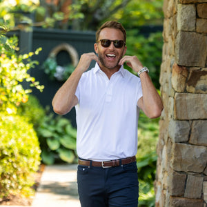A man in sunglasses and a white COLLARS AND CO - FEATHERWEIGHT FLEX POLO stands outdoors, smiling and holding his collar, with greenery and a stone wall in the background.