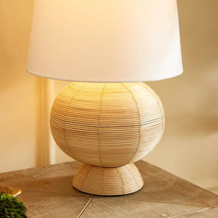 The NAPA HOME AND GARDEN Maye Rattan Hourglass Lamp with Shade features a wicker-style base and white shade, casting a warm glow on any wooden surface.