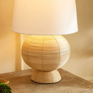 The NAPA HOME AND GARDEN Maye Rattan Hourglass Lamp with Shade features a wicker-style base and white shade, casting a warm glow on any wooden surface.