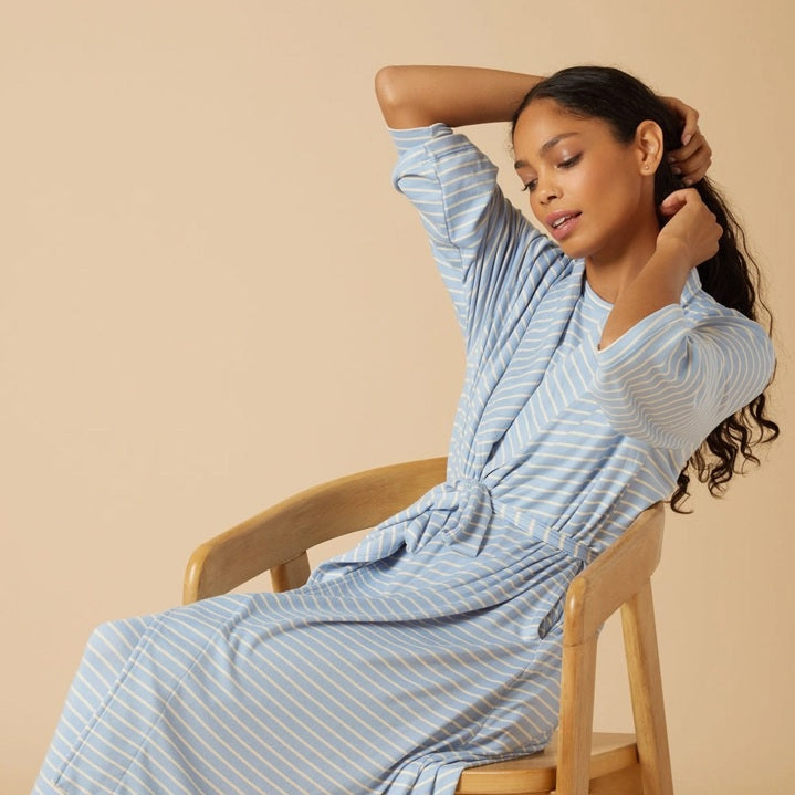 Wearing light blue and white striped pajamas, a person stands barefoot against a beige background, draped in the SOFTIES Dream Shawl Collar Robe in Sky Blue Stripe. This lightweight robe from SOFTIES adds an extra layer of silky smooth comfort to the cozy look.