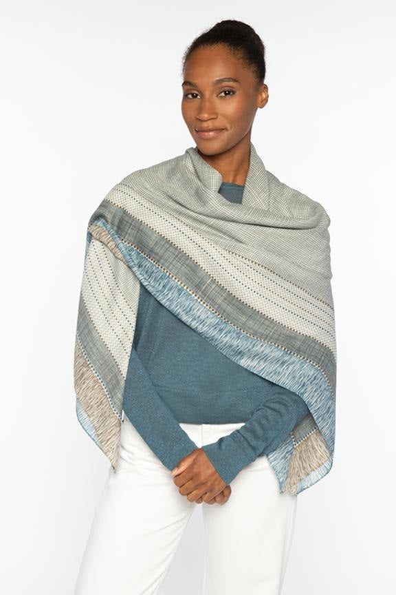 A woman in a blue sweater and white pants smiles softly with her hands folded, showcasing the KINROSS CHECKED OUT CASHMERE SCARF by KINROSS, its soft gray and blue checkered pattern standing out against a plain white background.