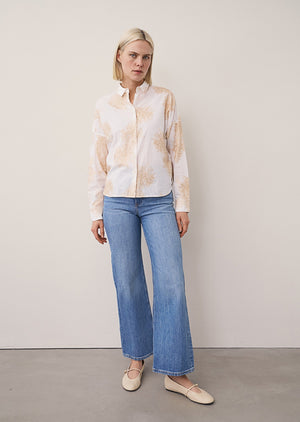 A close-up of the 0039 ITALY LUCY BLOUSE WITH EMBROIDERED FLOWERS, featuring detailed beige floral embroidery on pure cotton. A hand rests in the pocket, emphasizing the relaxed tailoring; the model’s face is out of frame.