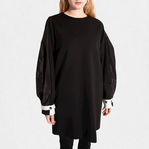 Dressed in the MEIMEIJ Knit and Taffeta Tunic/Dress, a versatile black piece with flowing sleeves and white cuffs, the ensemble is elegantly completed with black pants featuring subtle ankle slits, standing out against a plain white backdrop.