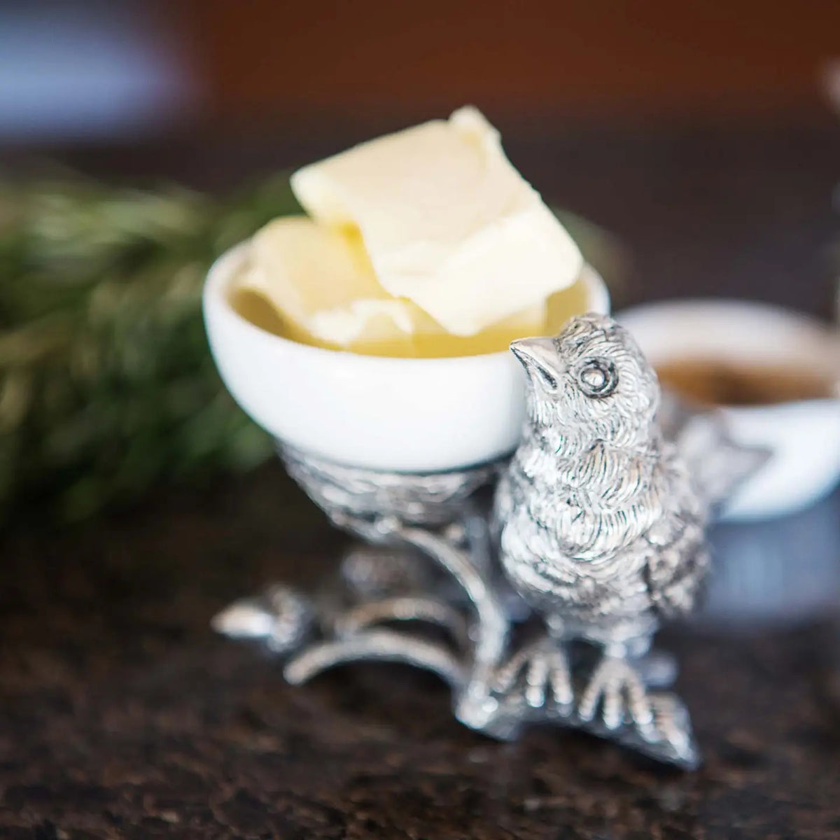 A small white bowl of butter sits on Vagabond House&#39;s Songbird Salt Cellar, showcasing intricate pewter designs with delicate feathers. The scene is set on a dark surface with blurred greenery in the background.