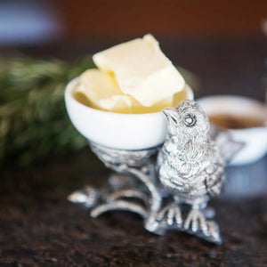 A small white bowl of butter sits on Vagabond House's Songbird Salt Cellar, showcasing intricate pewter designs with delicate feathers. The scene is set on a dark surface with blurred greenery in the background.