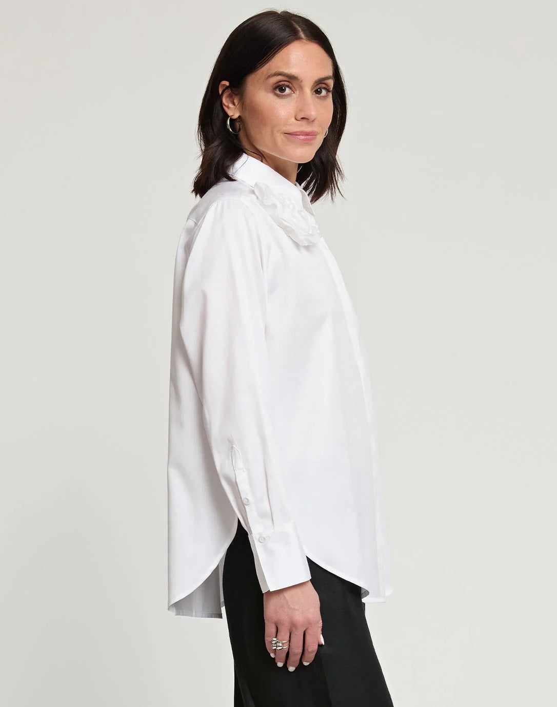 A person with short, brown hair is standing and facing away from the camera, wearing a HINSON WU - HALSEY LONG SLEEVE COTTON SATIN SHIRT WITH FLOWER. They have paired this black button-up shirt, made of a cotton and spandex blend, with light-colored pants. The background is plain and light-colored.