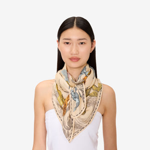 A person with long dark hair wears the INOUI EDITIONS Astrologie Scarf—a silk modal blend adorned with intricate zodiac signs—over a white strapless top, set against a plain white background.