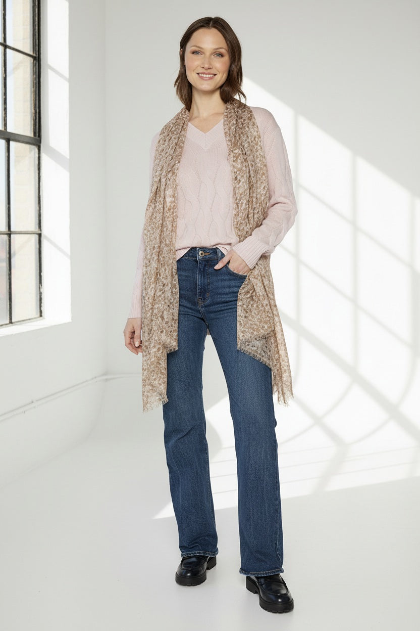A woman poses against a white background wearing a light pink sweater, blue jeans, black shoes, and the KINROSS CASHMERE - PEBBLE PRINT SCARF by KINROSS in a silk cashmere blend draped over her shoulders.