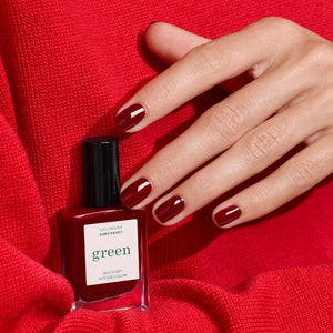 A hand with neatly manicured nails painted in a rich shade of dark red rests on red fabric beside a matching bottle of MANUCURIST INC’s GREEN™ Nail Polish in Dark Pansy, a non-toxic and vegan formula.