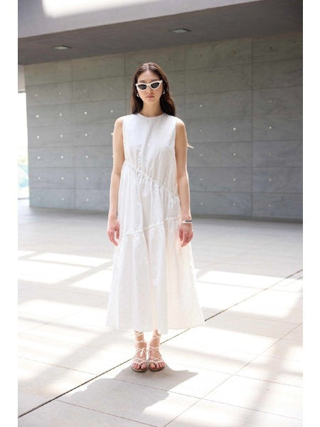 A woman in the ELEH - SLEEVELESS COTTON DRESS with white sunglasses and strappy sandals stands in a bright, modern space with large windows and a gray tiled wall, showcasing effortless resort style by ELEH.