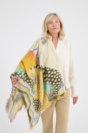A woman holds the STORIATIPIC MALI MAX SCARF IN BEIGE, a luxurious cotton-modal accessory with colorful abstract and botanical patterns, against a plain white background.