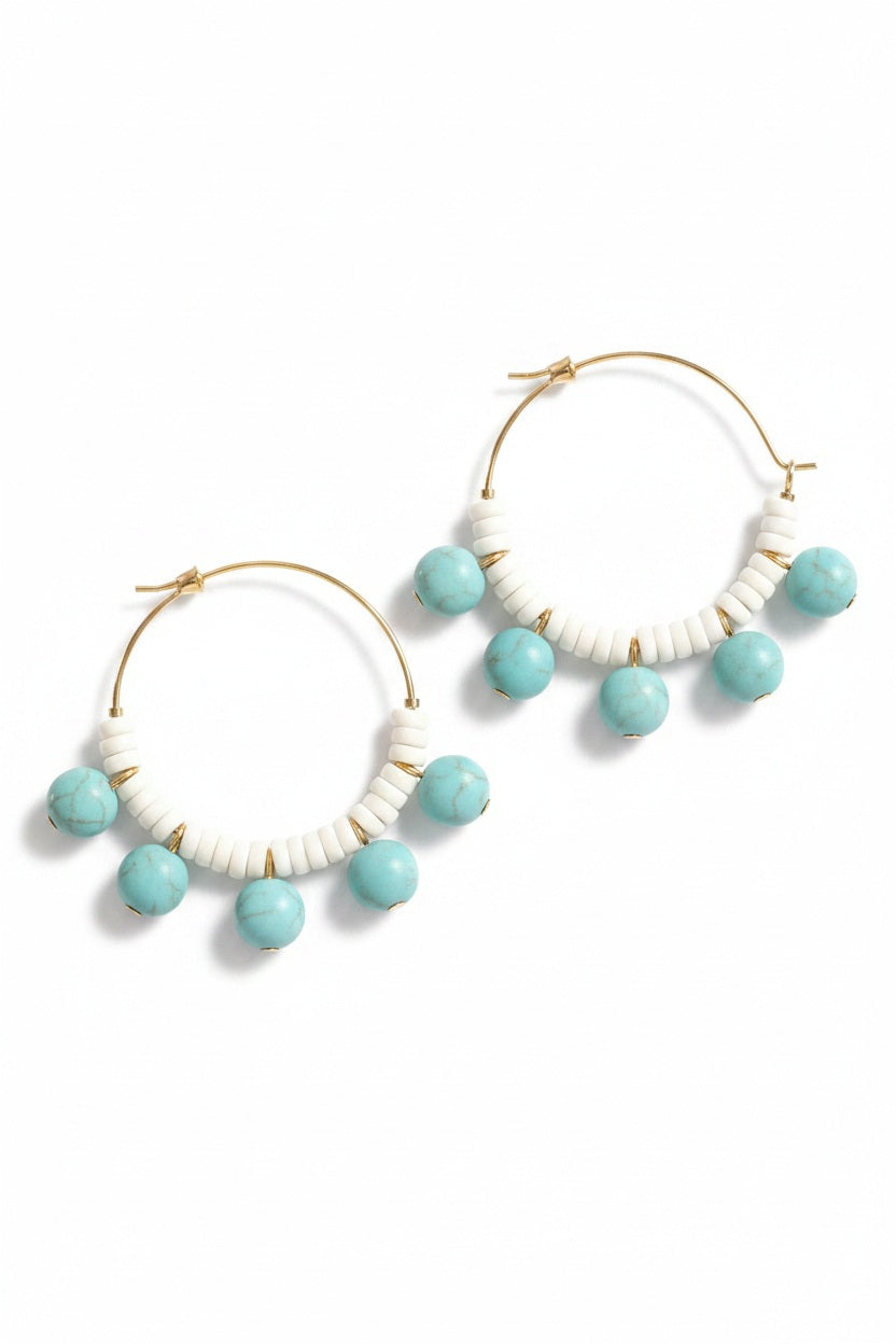 DAVID AUBREY - TUQUOISE AND WHITE HOOP EARRINGS by DAVID AUBREY, featuring gold plated hoops embellished with alternating white magnesite heishi beads and round turquoise glass beads, are displayed on a black hexagonal surface against a dark gray background.