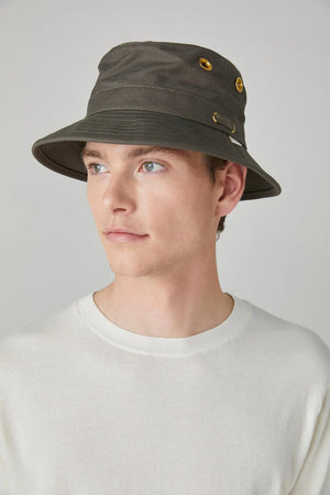 A young man in a white shirt is shown in profile wearing the TILLEY - ICONIC T1 BUCKET HAT in dark gray with golden eyelets. The UPF 50+ sun protection hat stands out against the plain, light gray background.