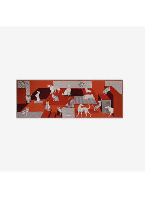 A person dressed in a cream outfit models the INOUI EDITIONS - LES CHERIS WOOL SCARF IN RED by INOUI—a 100% wool accessory made in India, featuring bold orange, red, and burgundy tones adorned with large white cat illustrations and geometric patterns—set against a plain background.