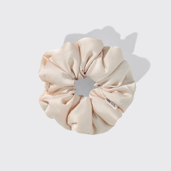 A package of KITSCH PILLOW SATIN SCRUNCHIES IN BLUSH sits behind four stacked scrunchies in beige, pink, cream, and champagne shades. The packaging displays a woman’s face and reads “Pillow Scrunchies in Satin” by KITSCH, showcasing this gentle, damage-free hair accessory ideal for soft styling.