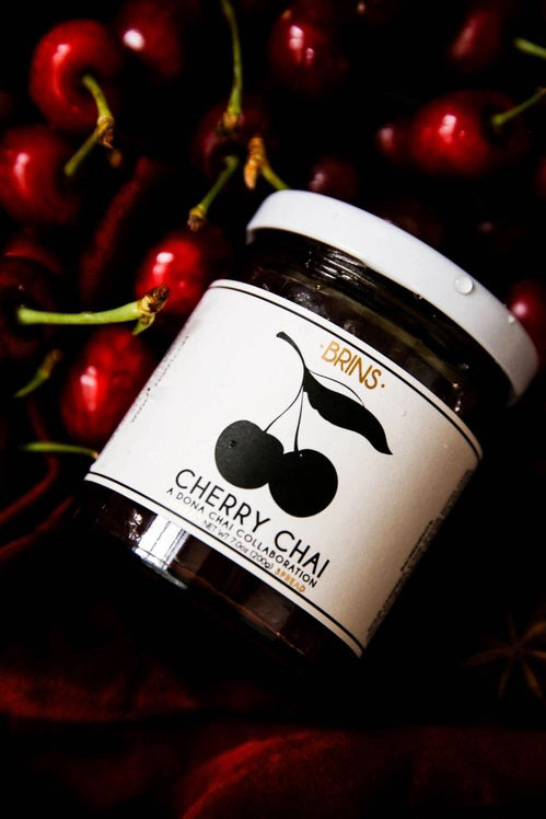A glass jar of BRINS - CHERRY CHAI SPREAD & PRESERVE features a black lid and a label adorned with an illustration of two cherries alongside flavor details and collaboration notes. Made by BRINS with locally sourced New York sour cherries, this preserve is expertly infused with aromatic chai spices for a distinctive and flavorful experience.