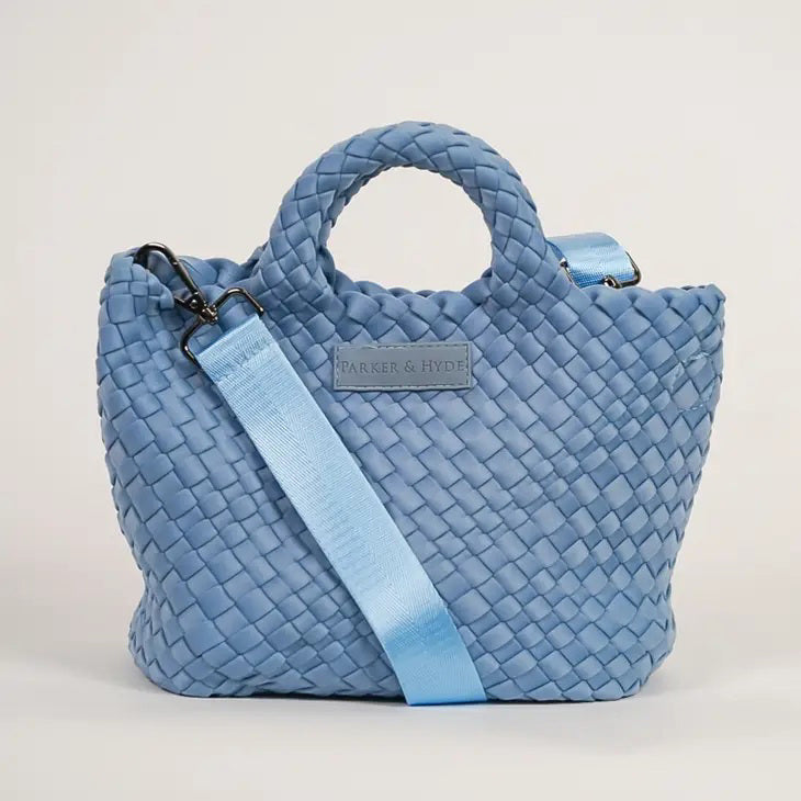 A woman with long brown hair, in a sleeveless cream knit top and blue jeans, holds a PARKER AND HYDE - MINI WOVEN TOTE BAG. The hand-woven tote by PARKER &amp; HYDE features a subtle checkered pattern with light blue accents.