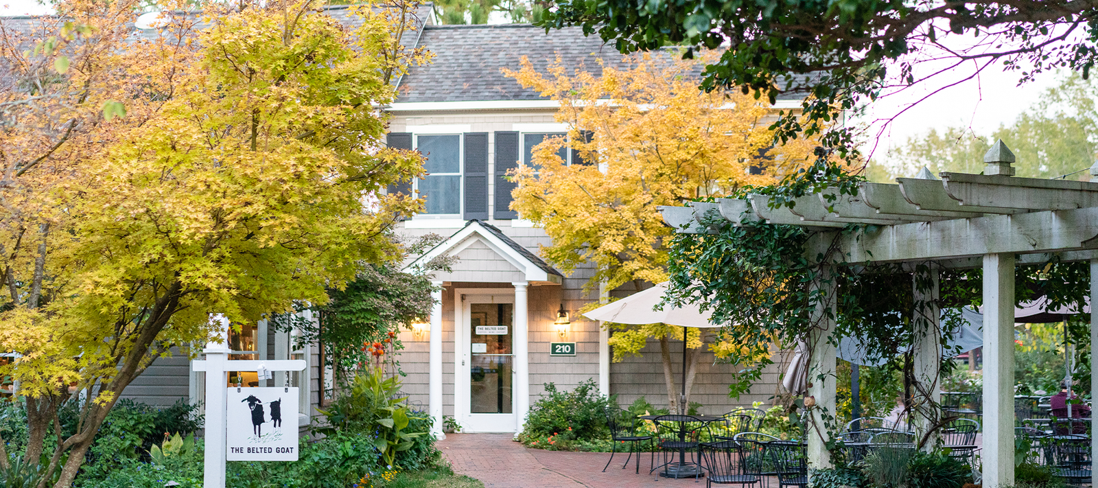 A charming house with beige siding and white trim, surrounded by autumn trees with yellow leaves. A white sign is in front, and a pergola with outdoor seating and tables is on the right. A brick path leads to the entrance.