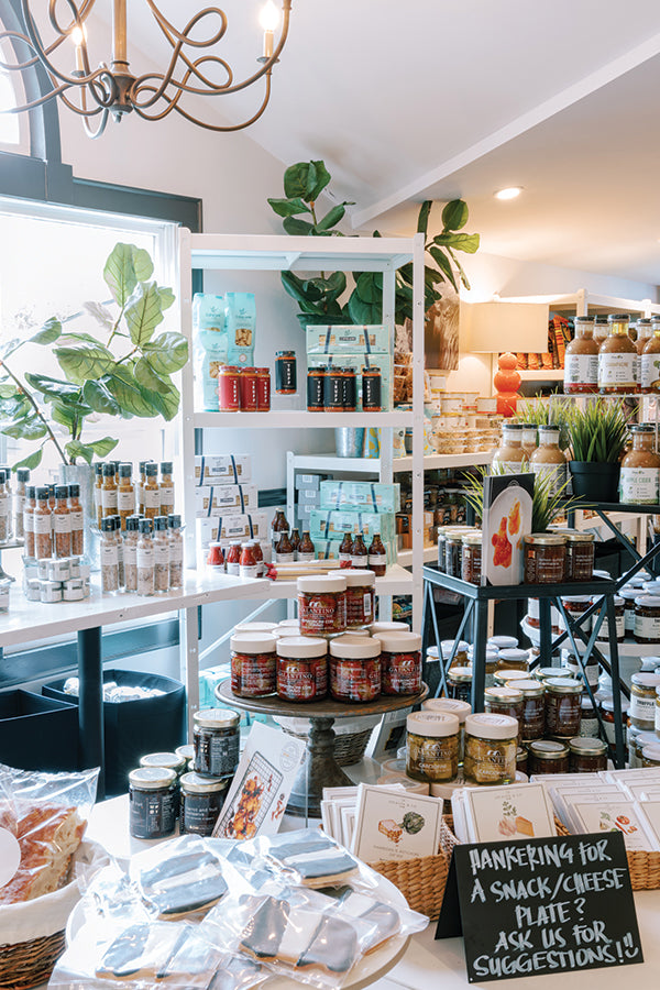 A bright, cozy shop interior with shelves displaying various jars of jams, sauces, and condiments. Green plants decorate the space and a sign suggests asking staff for snack or cheese plate recommendations.